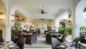 A spacious indoor dining area with high ceilings, arched columns, and wicker furniture. Tables are set with plates, cutlery, and glassware. - Swahili Beach Resort (Diani Beach)