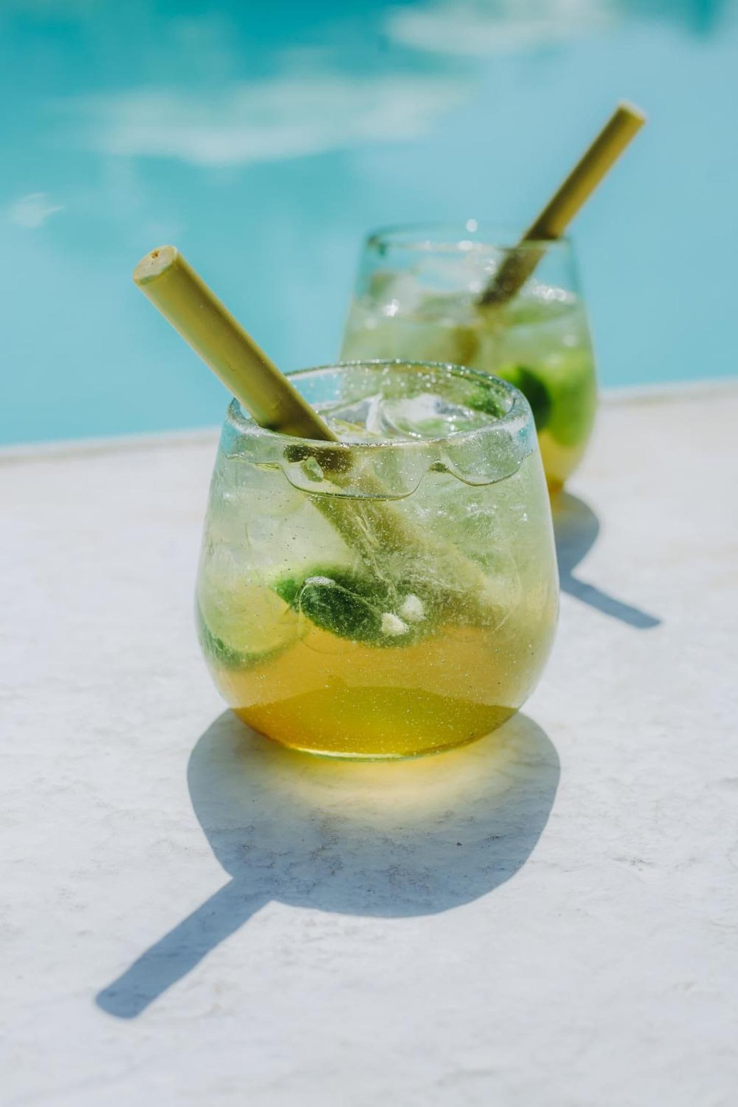Two glasses of a yellow beverage with mint leaves and a straw, placed on a surface with a pool in the background.
