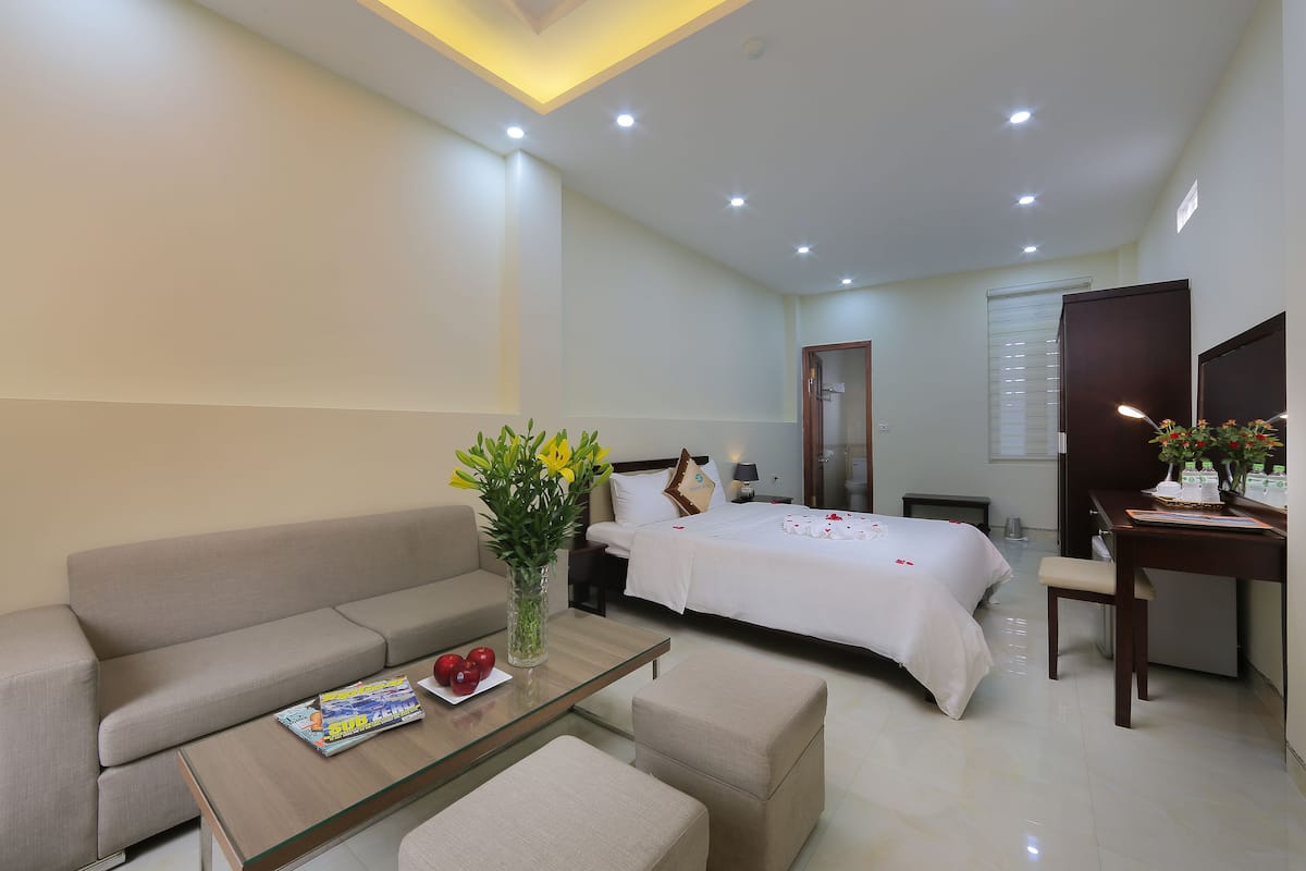 A modern hotel room with a sofa, a glass-top coffee table, a bed with white bedding, a desk with a chair, and a vase with flowers.