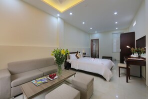 A modern hotel room with a sofa, a glass-top coffee table, a bed with white bedding, a desk with a chair, and a vase with flowers. - Spoon Hotel (Hanoi)