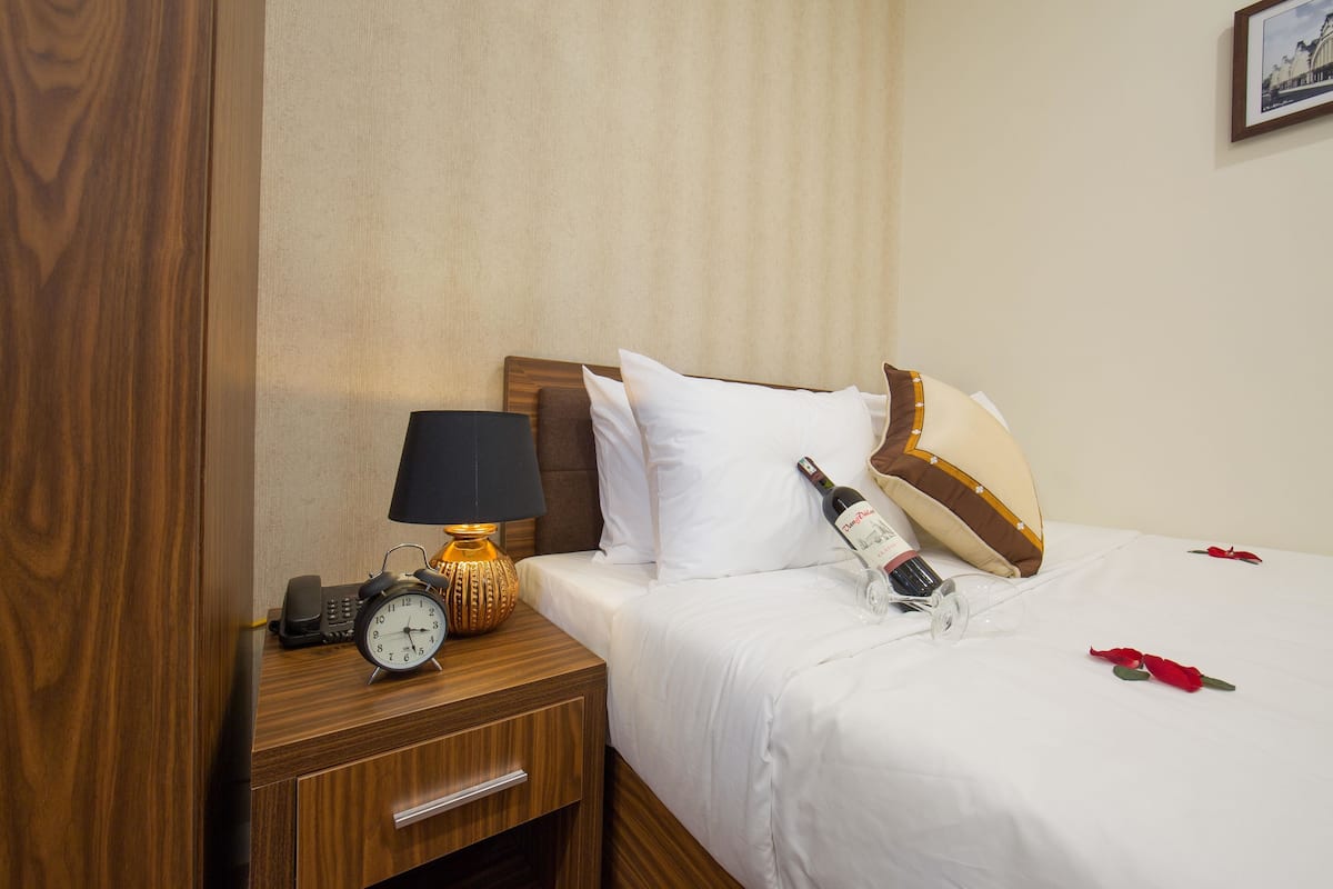 A neatly made hotel room with a wooden nightstand, a clock, a lamp, a bed with white bedding, and a bottle of wine.