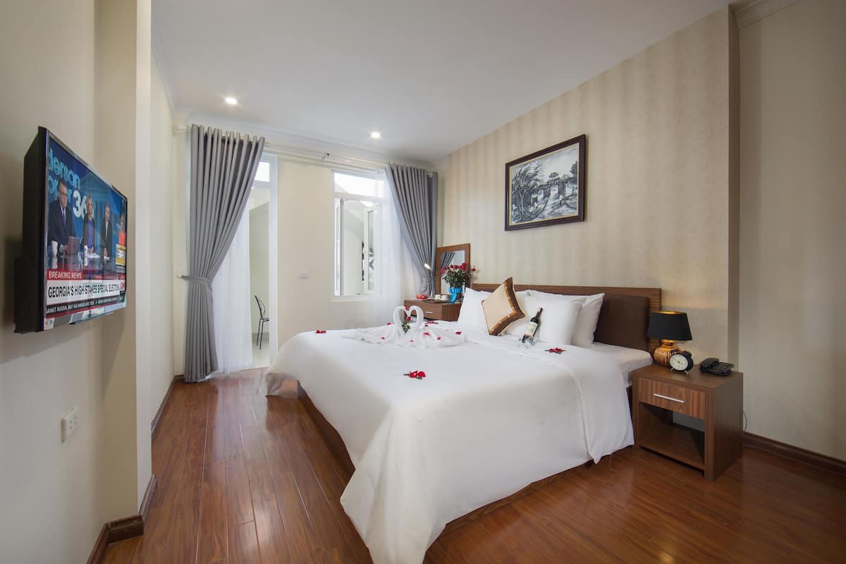A hotel room with a large bed, a bedside table with a lamp, a TV mounted on the wall, and a painting on the wall.