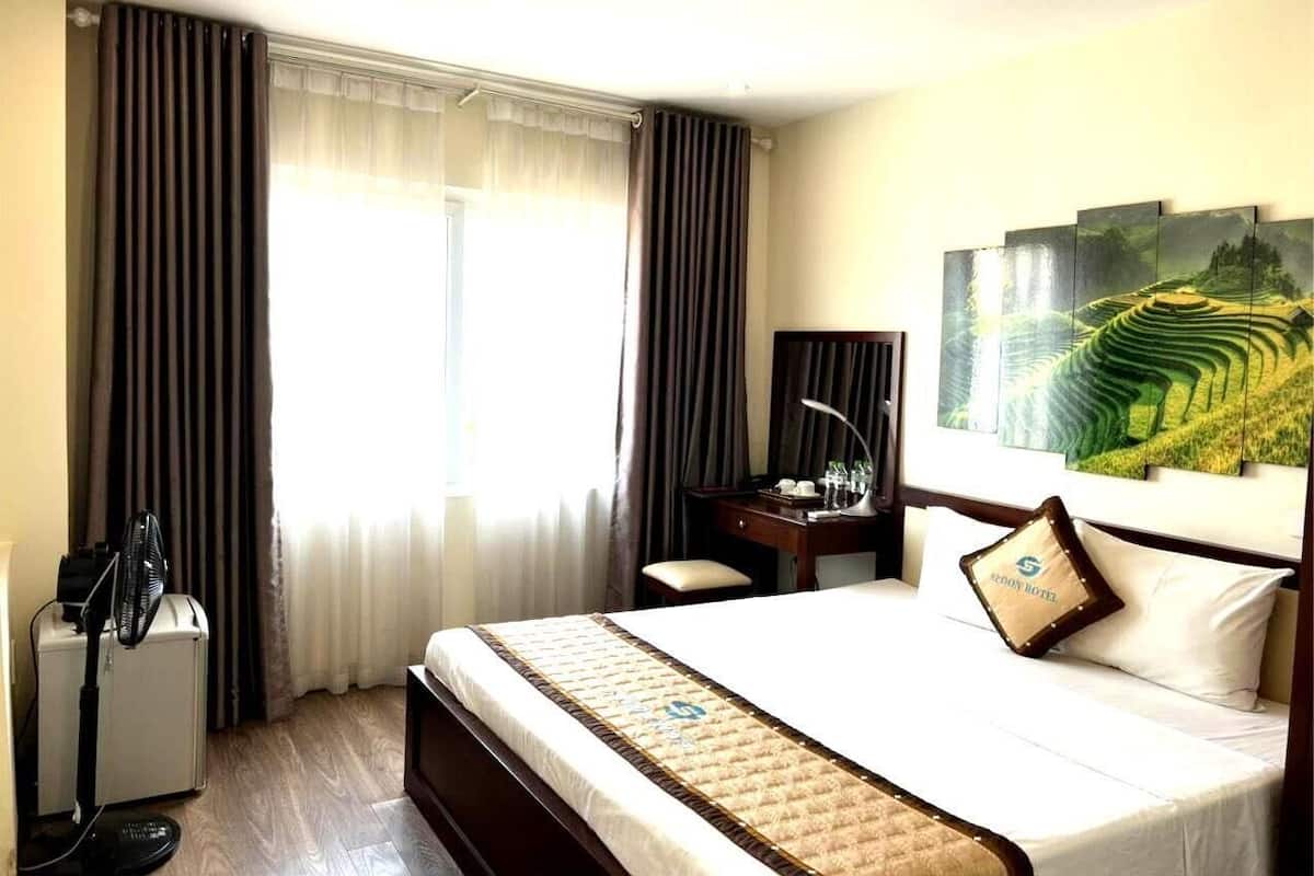 A hotel room with a large bed, a desk, a chair, a standing fan, and a painting on the wall.