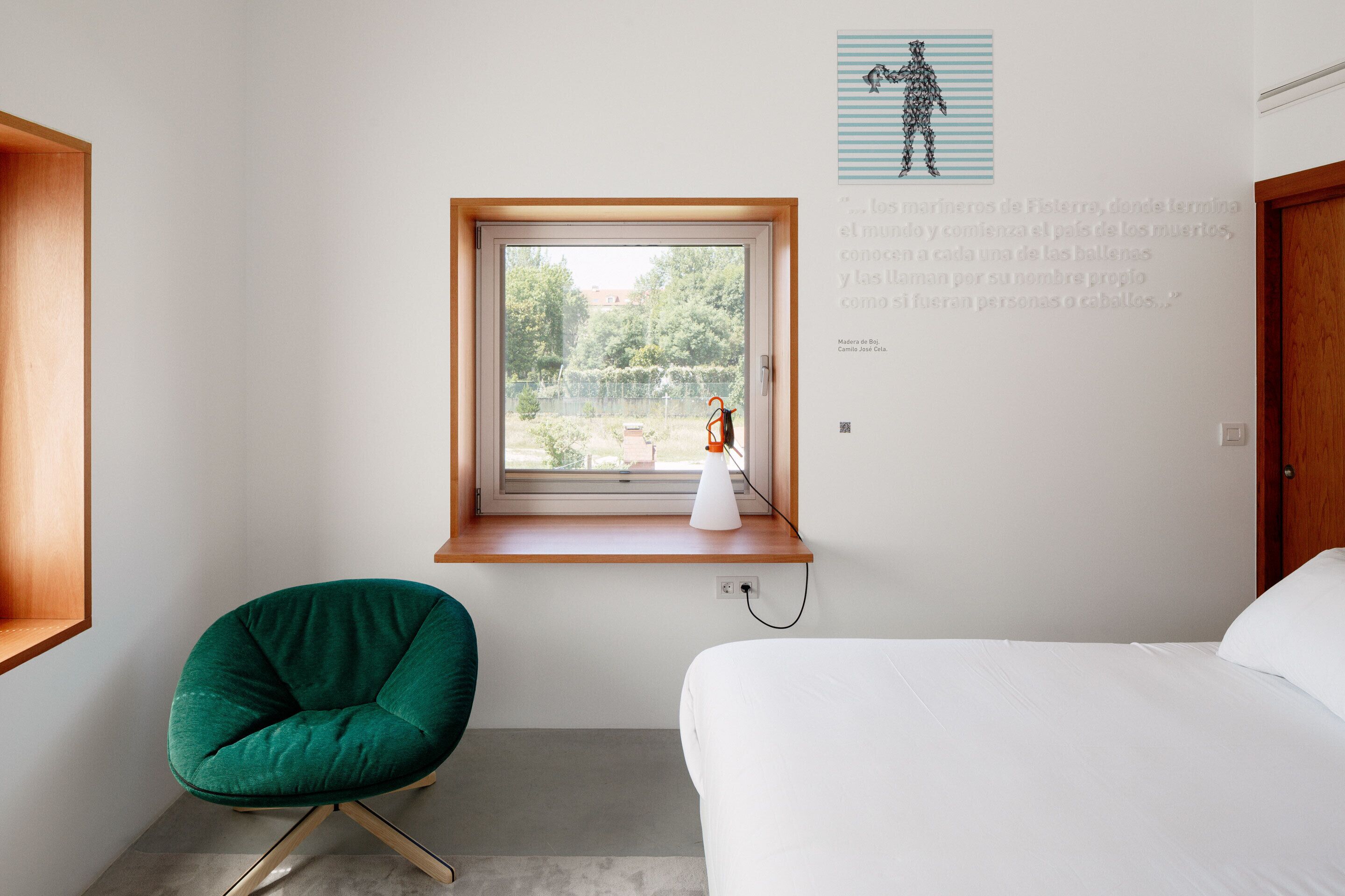 Un dormitorio con una cama, una silla verde, un ventanal con vistas a zonas verdes y una pared con un texto y una pequeña escultura.