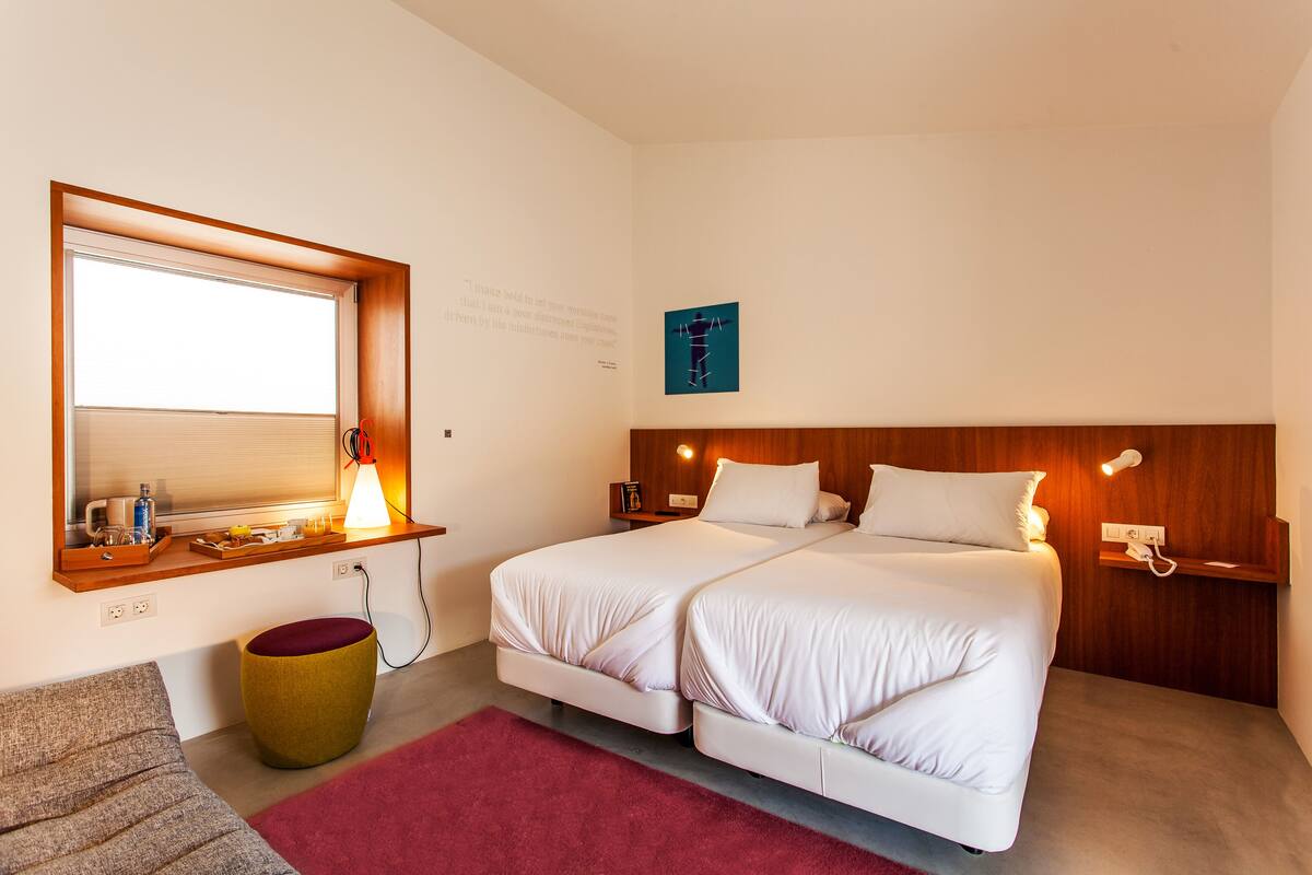 Una habitación de hotel moderna con dos camas, un cabecero de madera, una mesita de noche con una lámpara, una ventana con persianas y un taburete pequeño.