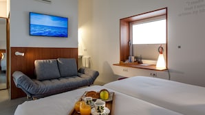 Ein Hotelzimmer mit Bett, Sofa, Frühstückswagen mit Croissants, Saft und Obst, einem an der Wand befestigten Fernseher und einem Fenster mit Meerblick.