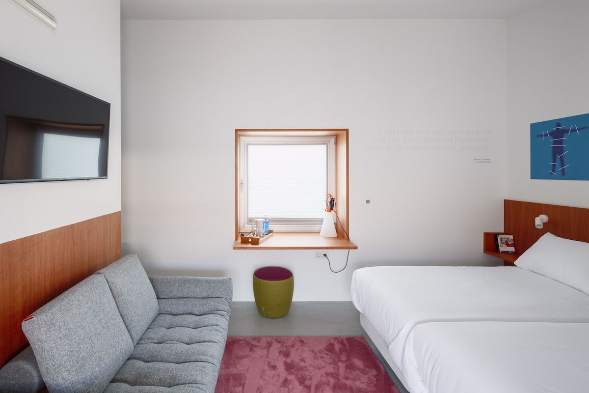 Habitación de hotel con cama, sofá, una ventana pequeña y un estante fijado a la pared con una lámpara y un libro.