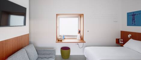 Habitación de hotel con cama, sofá, una ventana pequeña y un estante fijado a la pared con una lámpara y un libro.