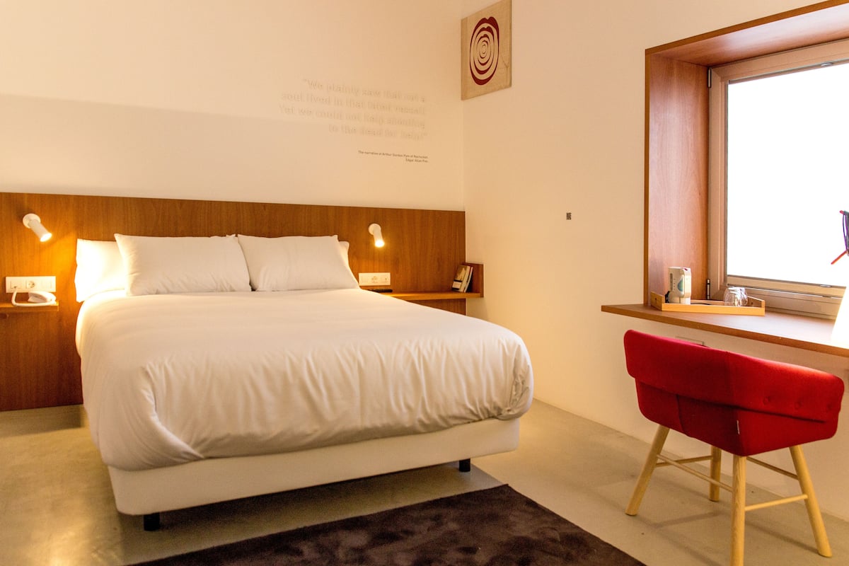 Una habitación de hotel ordenada con una cama grande, mesitas de noche, un escritorio con una silla y una ventana.
