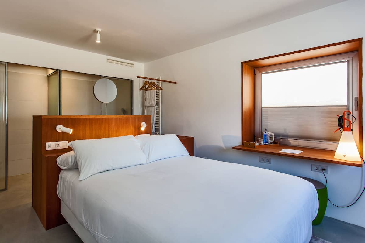 Una habitación de hotel moderna con un cabecero de madera, una cama grande con ropa blanca, una mesita de noche con lámpara, una ventana con persianas y un espejo en la pared.