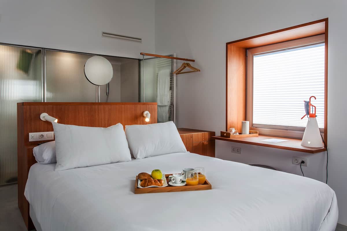 Una cama bien tendida con una bandeja de desayuno, una mesita de noche con lámpara y una ventana con persianas.
