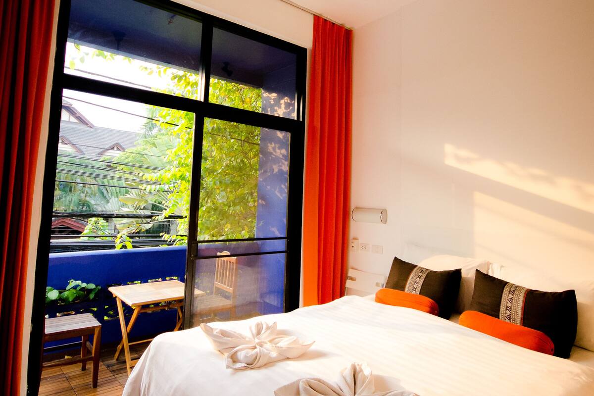 A hotel room with a bed, a ceiling fan, a window with curtains, and a view of greenery outside.