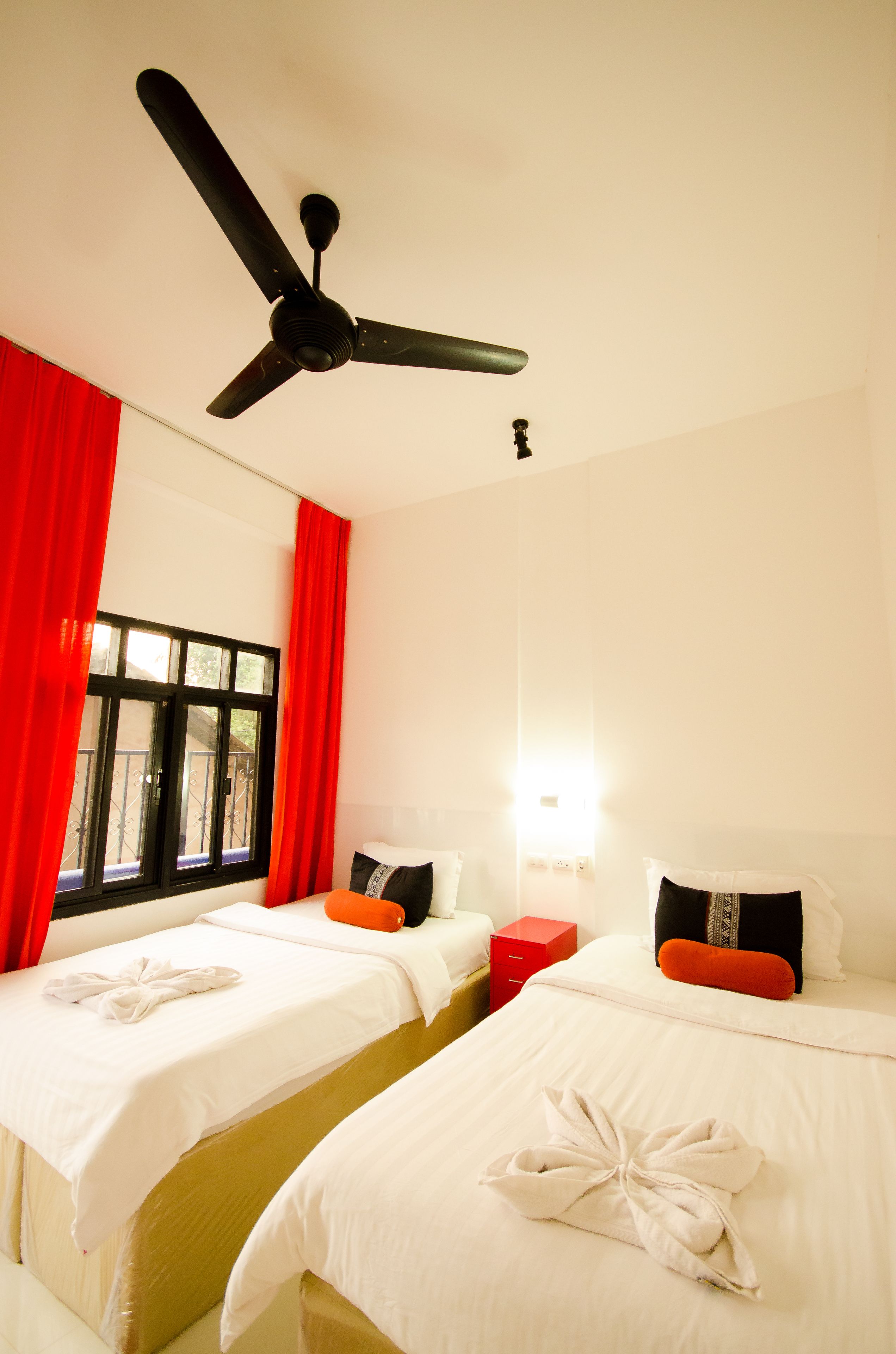 Une chambre d’hôtel avec deux lits, un ventilateur de plafond, une fenêtre avec des rideaux rouges et une table de chevet avec une lampe.