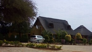 A swimming pool with a tiled edge, a paved area with a car, and a building with a thatched roof.