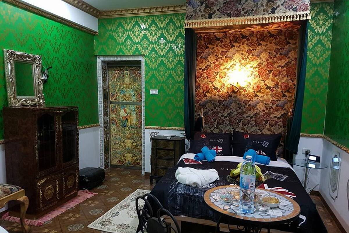 A room with green patterned wallpaper, a bed with a floral headboard, a small table with a bottle and glasses, and a mirror on the wall.