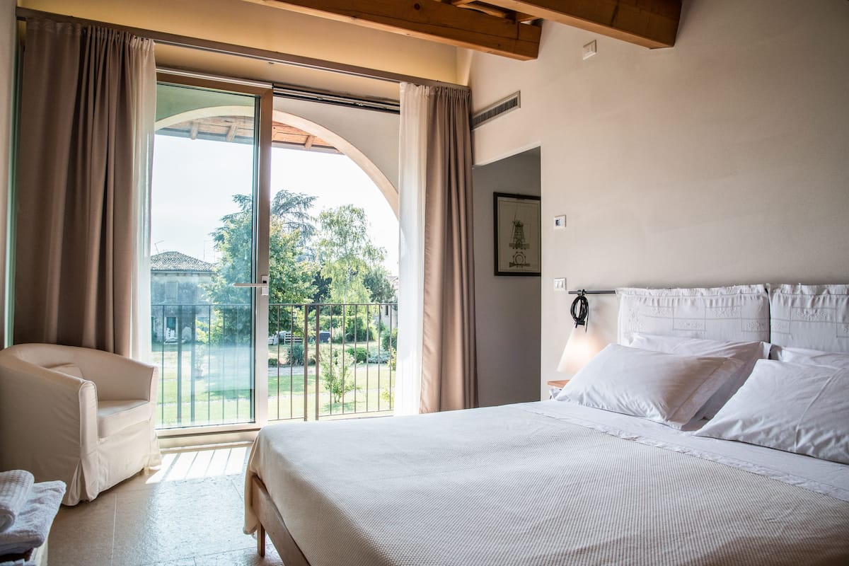 Un dormitorio con una cama grande, una silla y vistas a un jardín por un ventanal corredero.