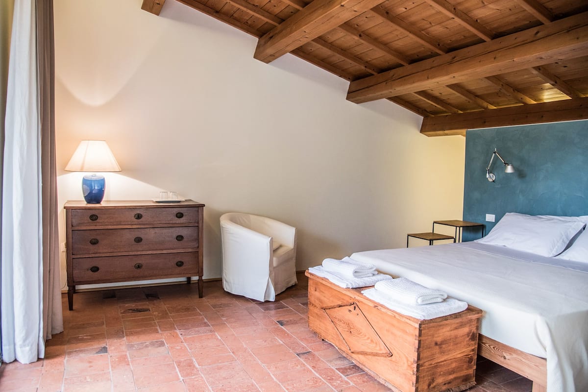 Un dormitorio con techo de madera, una cama con ropa de cama blanca, un aparador de madera, una silla blanca y una mesita de noche con lámpara.