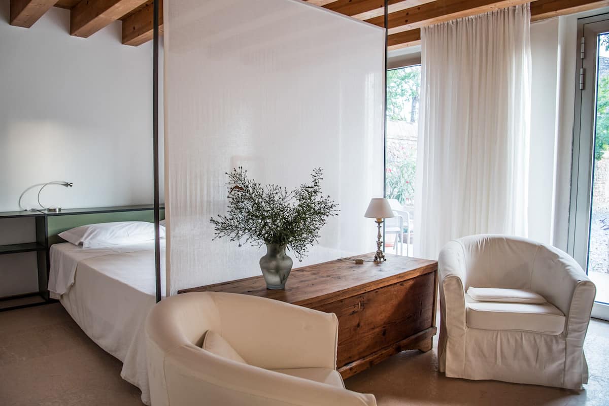 Un dormitorio con cama, mesita de noche, lámpara, silla y ventana con cortinas.