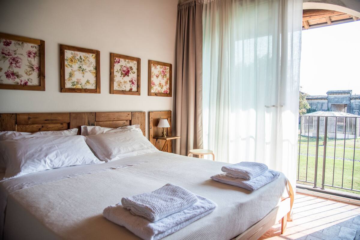 Una cama grande con sábanas blancas, cabecera de madera y vistas a un patio a través de un balcón con barandilla de metal.