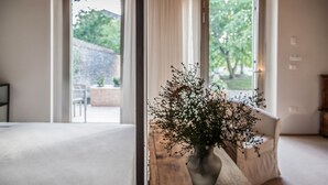 A room with a wooden table, a vase with flowers, and a view through a window to an outdoor area with a stone wall and greenery. - Agriturismo Corte Ruffoni (Zevio)