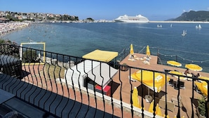 Une terrasse au bord de l’eau, agrémentée de parasols jaunes, avec vue sur la mer et un navire de croisière à l’horizon.
