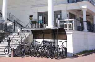 Zona techada para estacionamiento de bicicletas con varias bicicletas, una escalera y una entrada de edificio con columnas y un letrero.