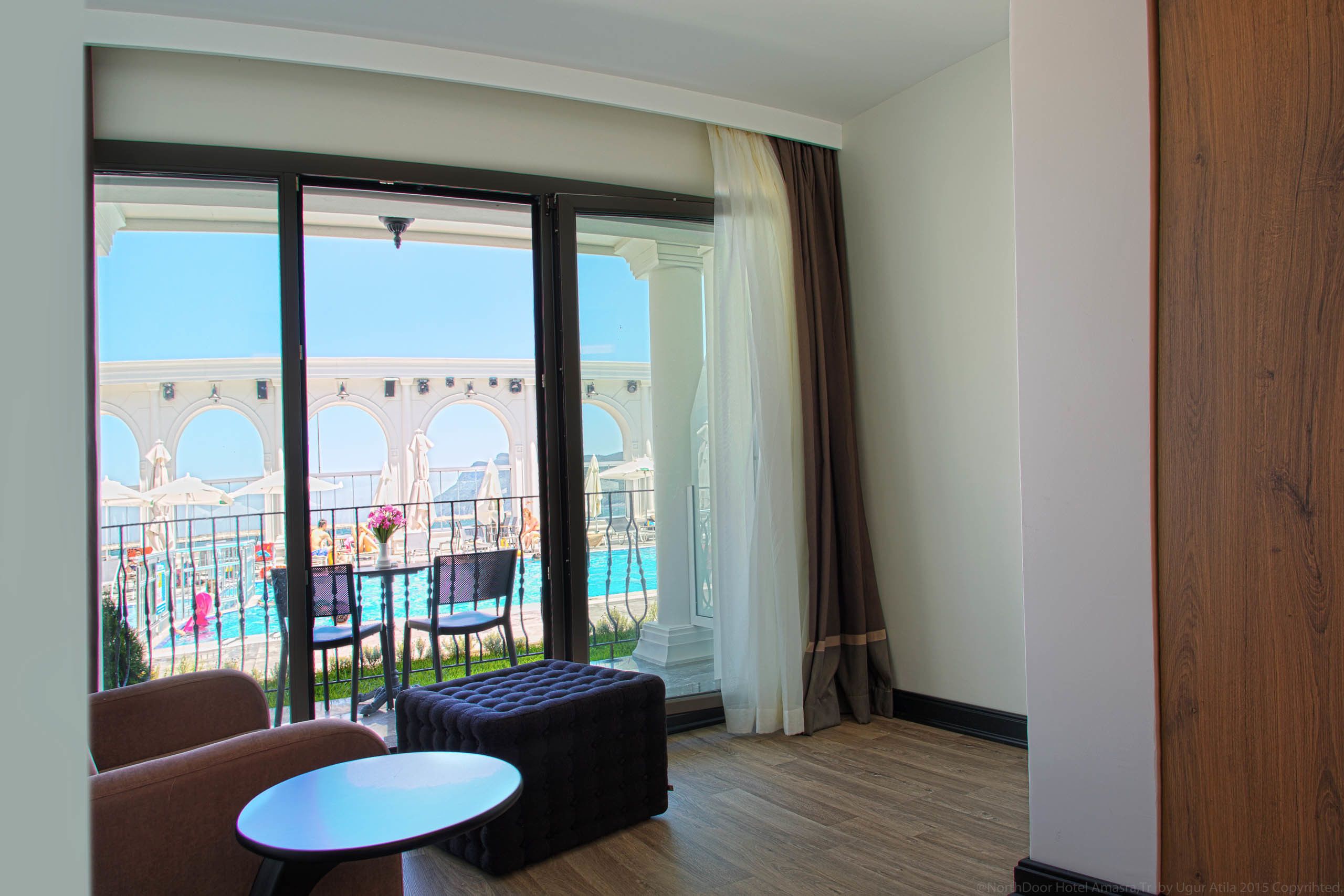 A hotel room with a large window, a small round table, a cushioned bench, and a view of a pool and building with arches.