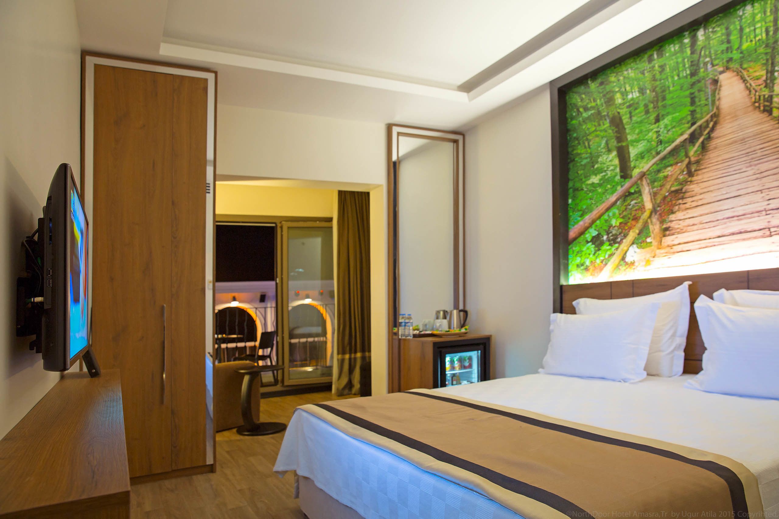 A hotel room with a bed, a TV, a mirror, and a painting of a forest path.