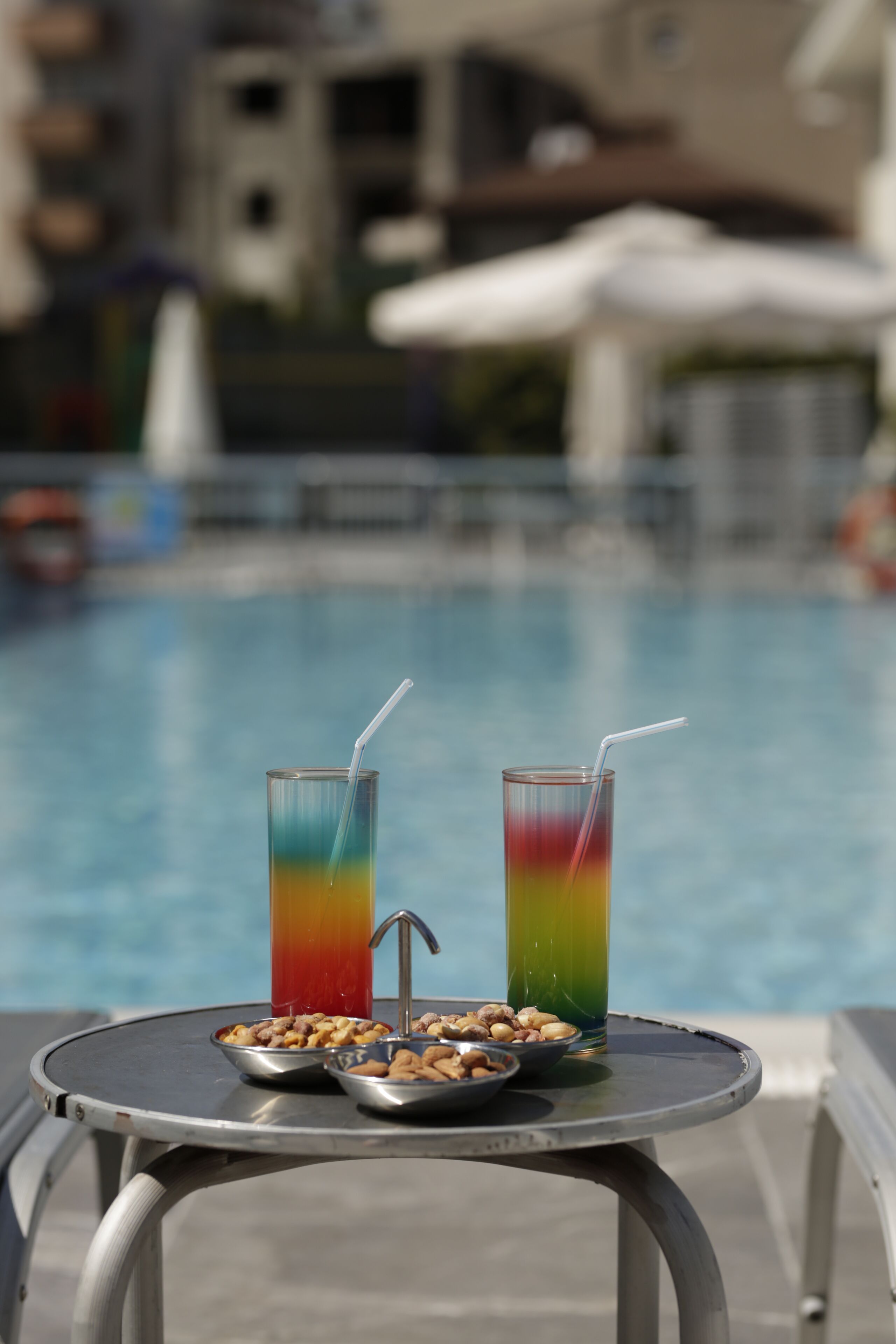 A table with two colorful drinks and bowls of nuts by a poolside.
