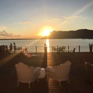 Terraza de madera con sillas y mesas blancas, con vista a un cuerpo de agua durante el atardecer.