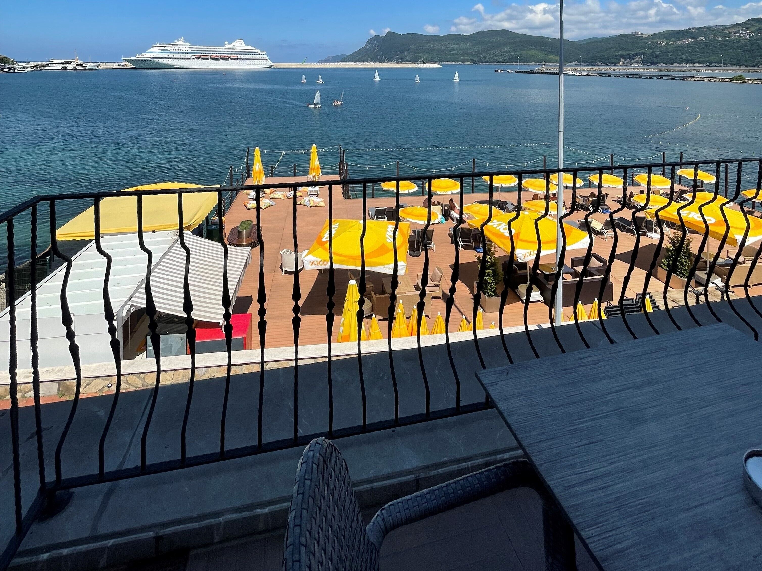 A deck with yellow umbrellas and seating, overlooking a marina with sailboats and a cruise ship.