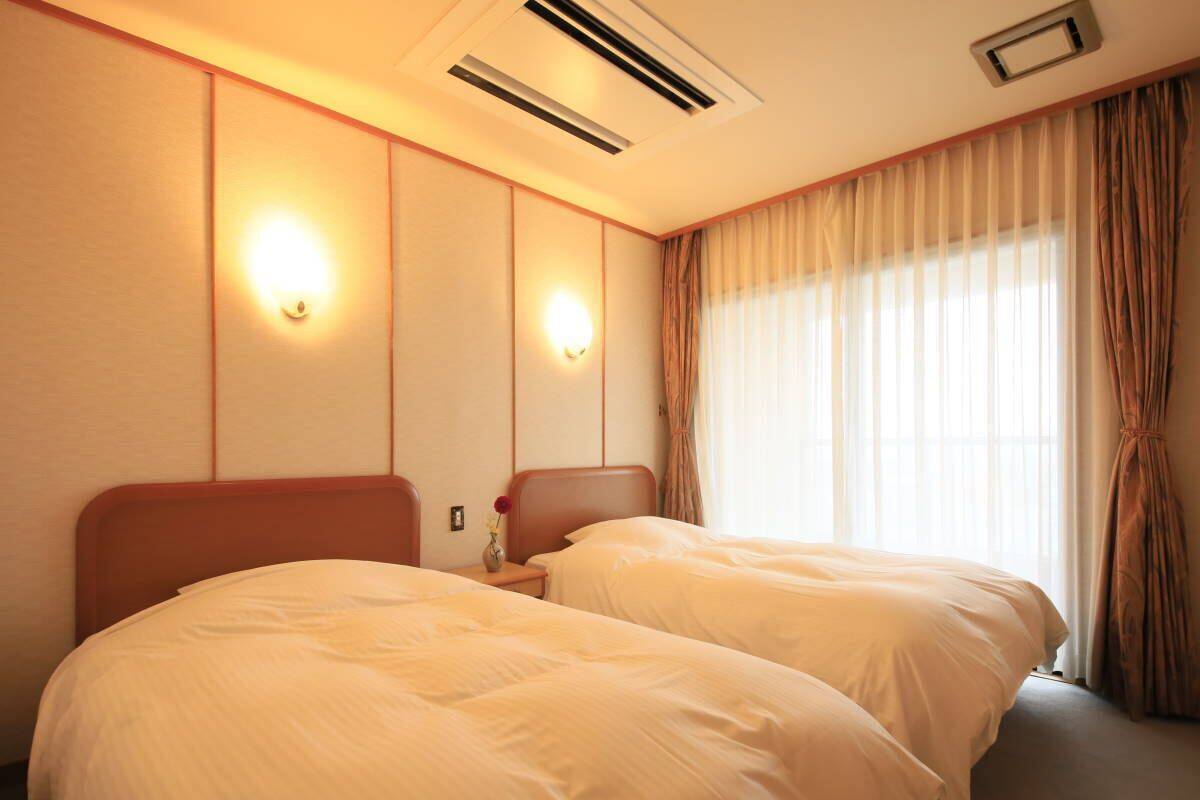 A hotel room with two beds, a nightstand, a wall-mounted lamp, and a window with curtains.