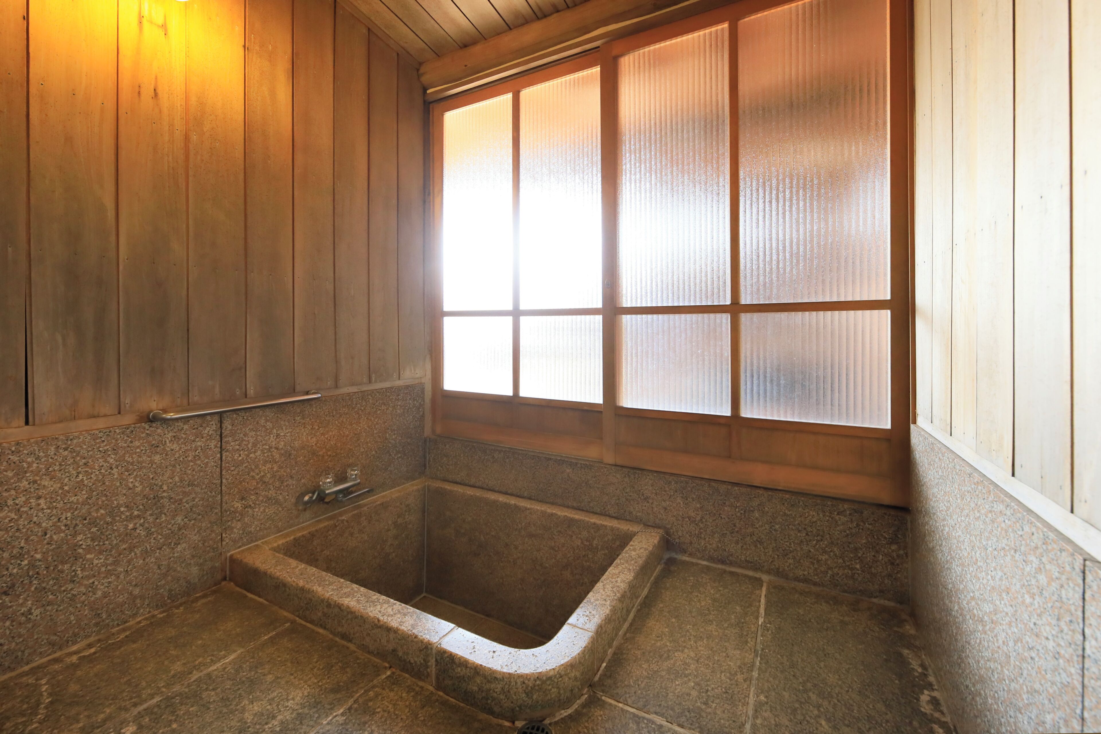 Une salle de bain de style japonais traditionnel, avec une baignoire en pierre, un lambris en bois et une fenêtre en verre dépoli.