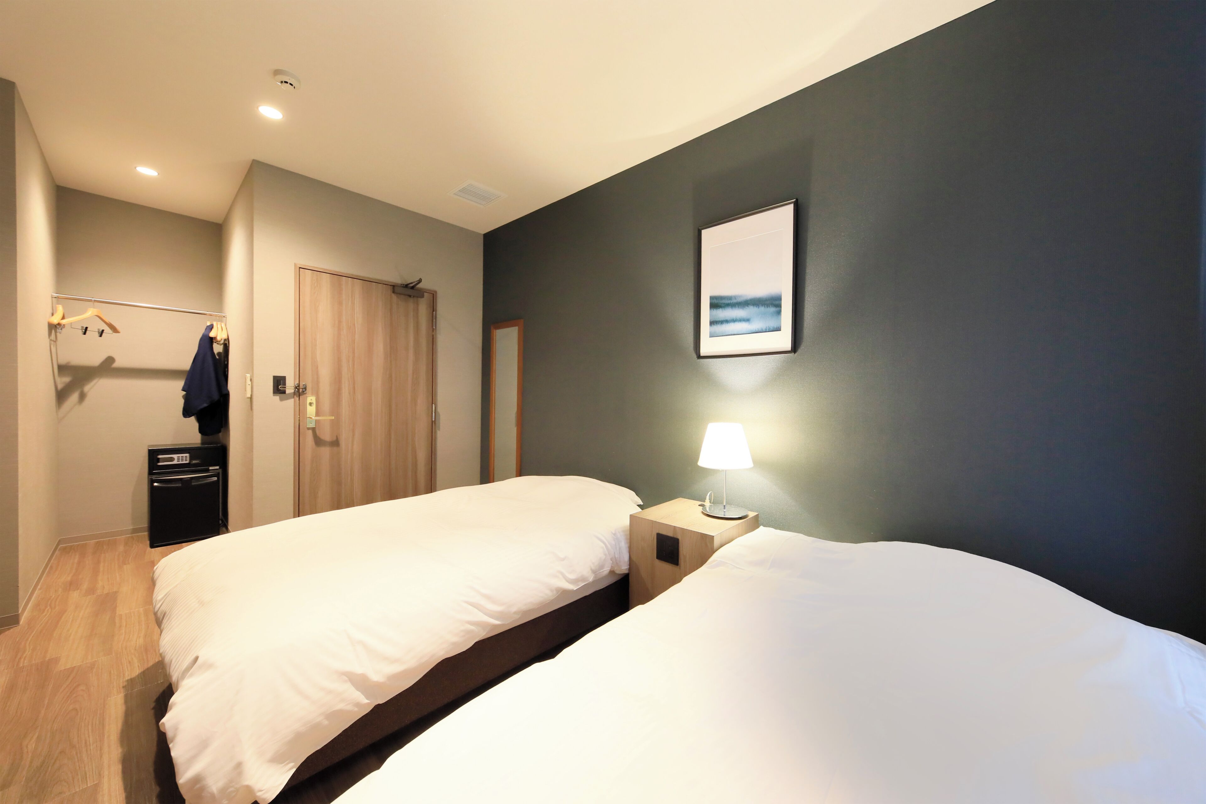 A hotel room with two beds, a bedside table with a lamp, a coat rack, and a door.