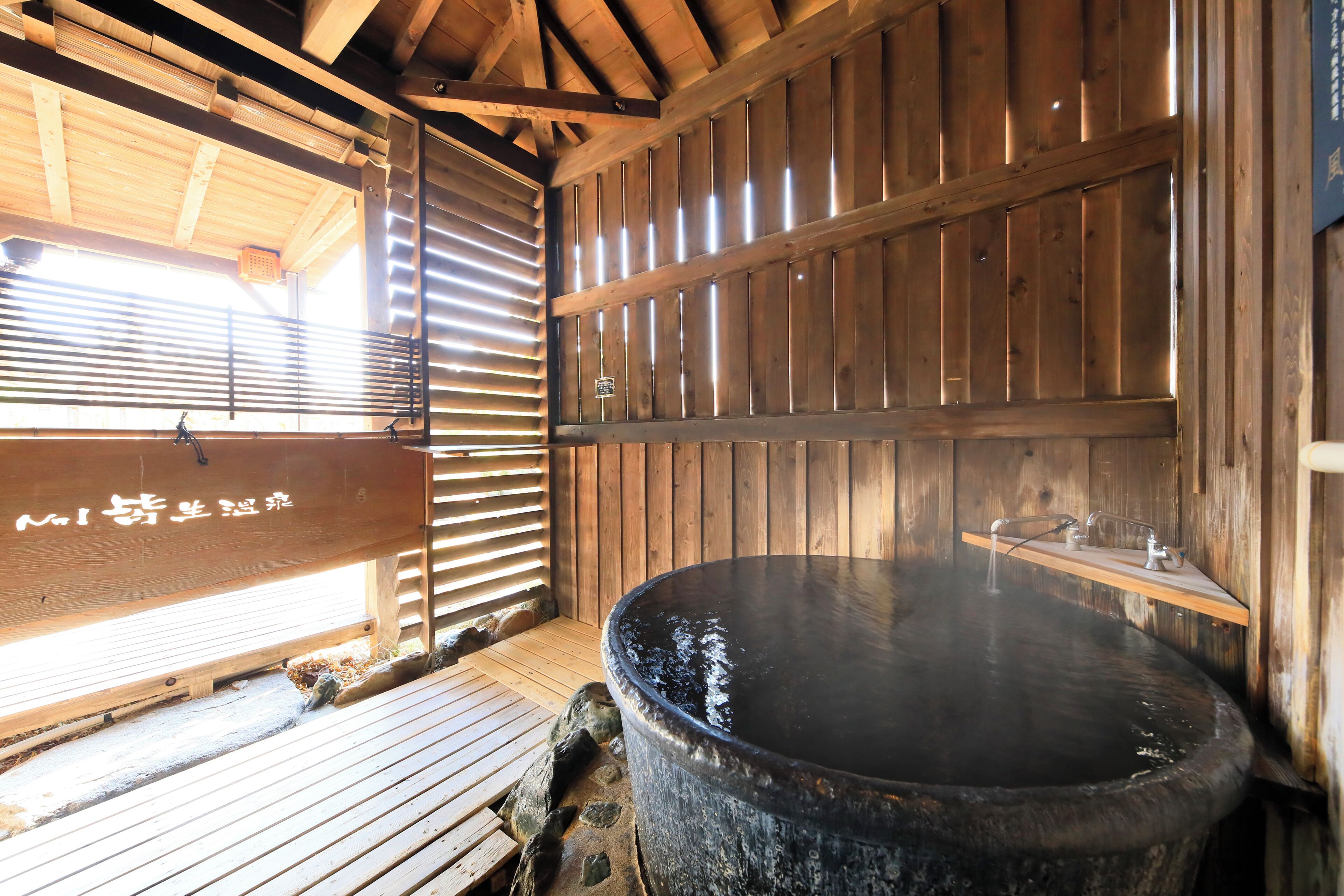 A traditional wooden bathhouse with a large stone hot tub, wooden benches, and a wooden ceiling structure.