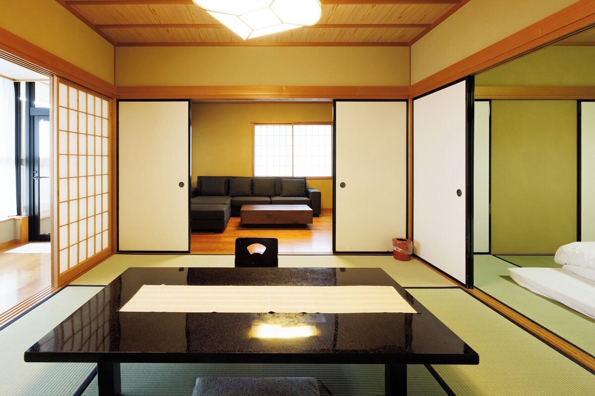 A traditional Japanese room with tatami flooring, a low table, a sofa, sliding doors, and a view of a cityscape.