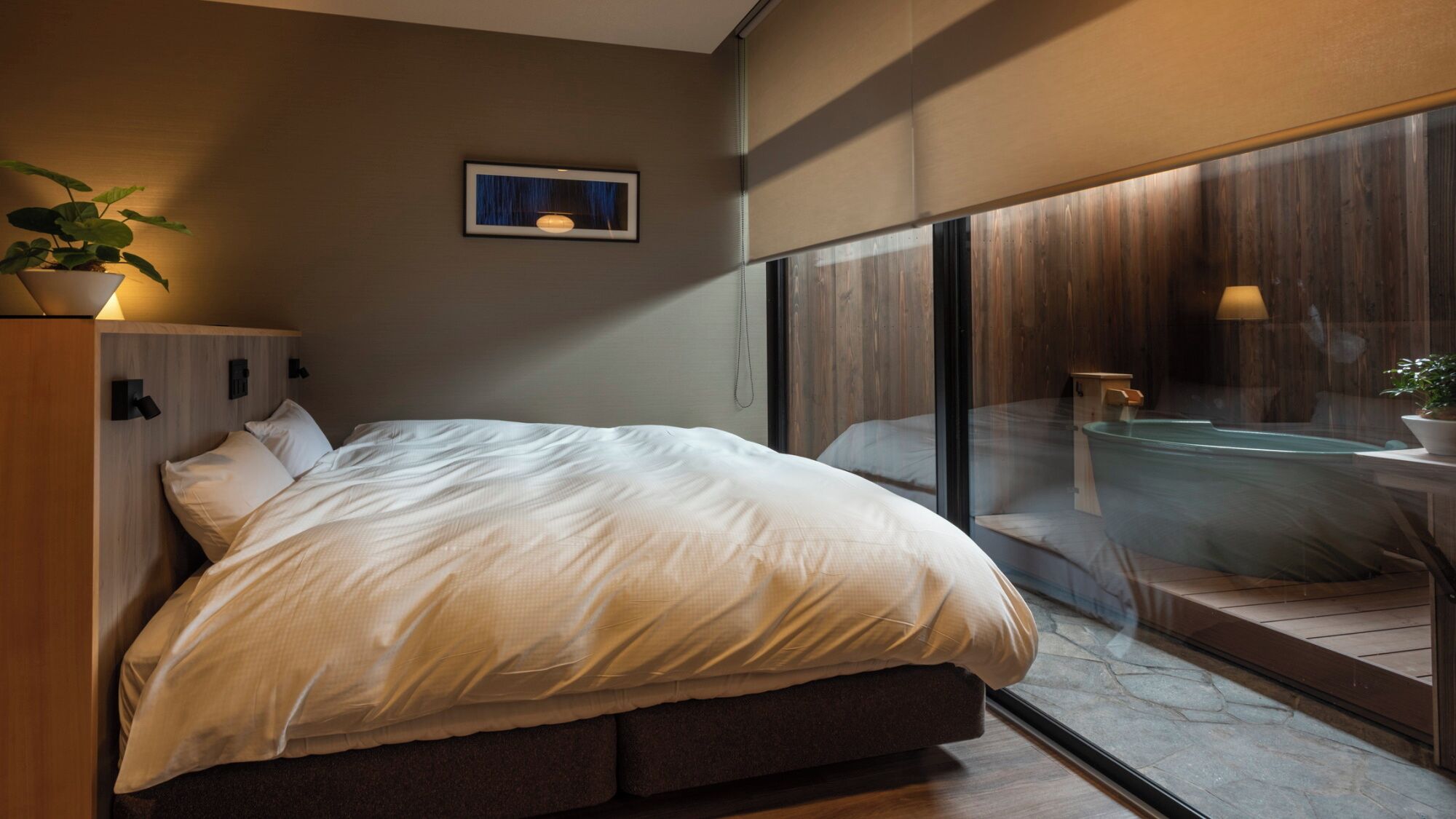 A modern bedroom with a large bed, a bedside table, a potted plant, and a view of a spa area through a glass wall.