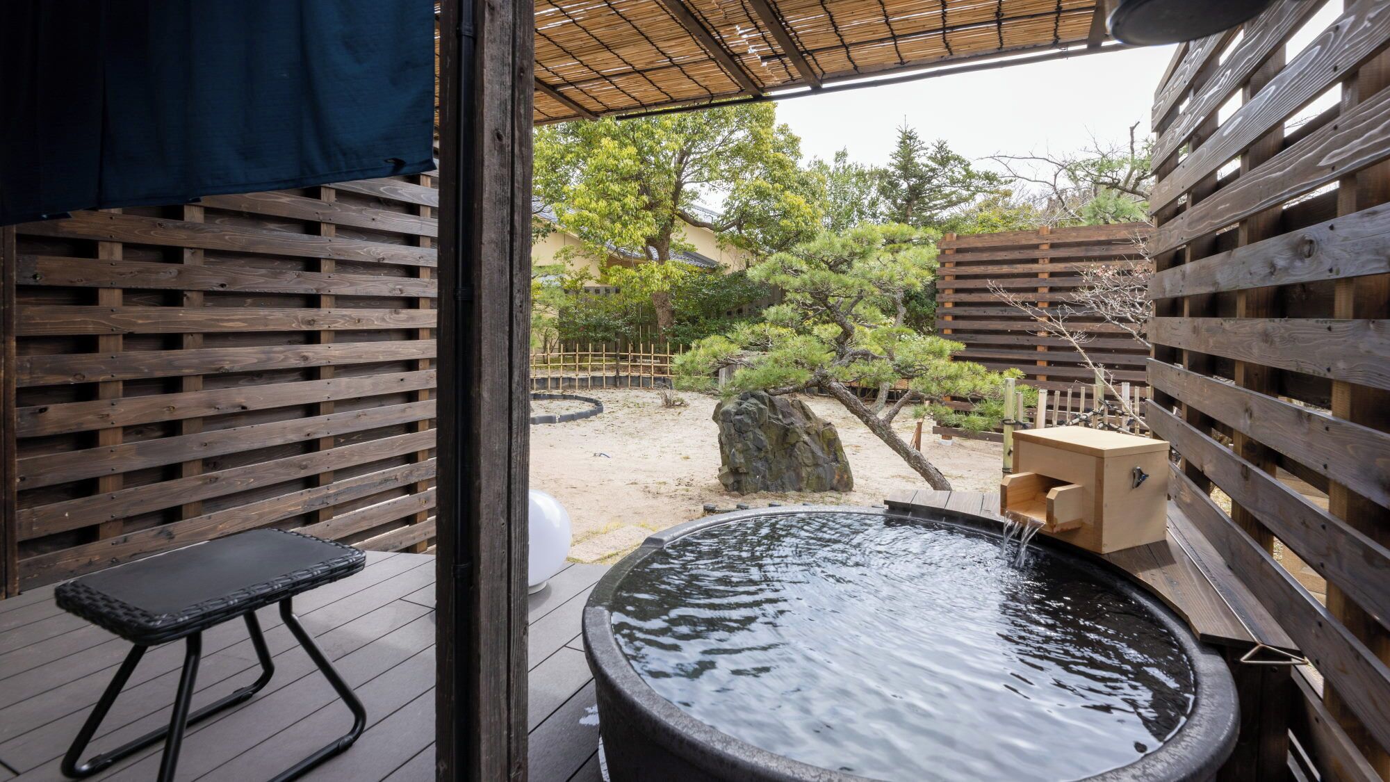 Zona de aguas termales con una terraza de madera, una bañera de piedra caliente y vistas a un jardín con árboles y un edificio al fondo.