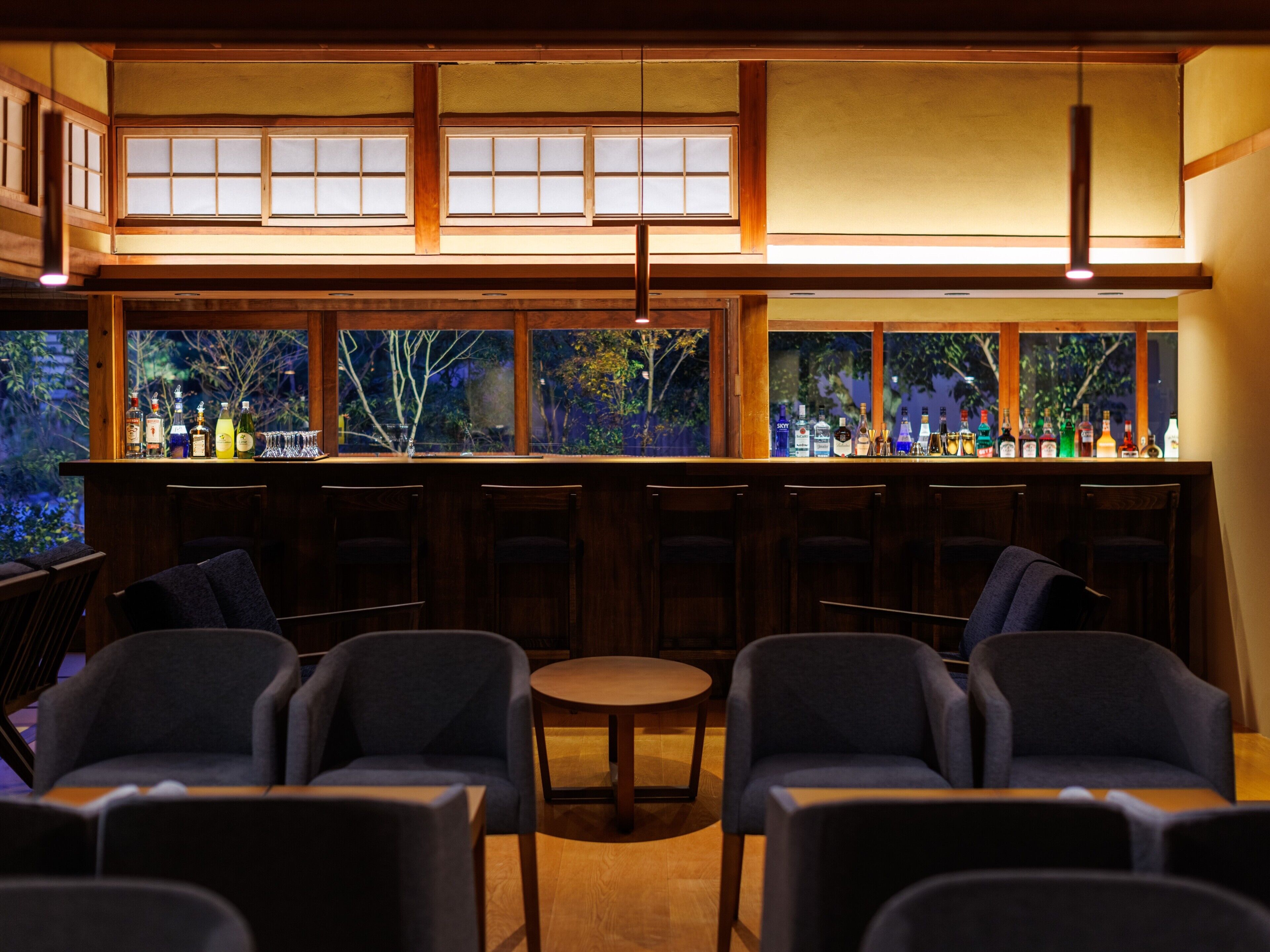A bar area with wooden furniture, a bar counter with bottles, and a view of trees through large windows.