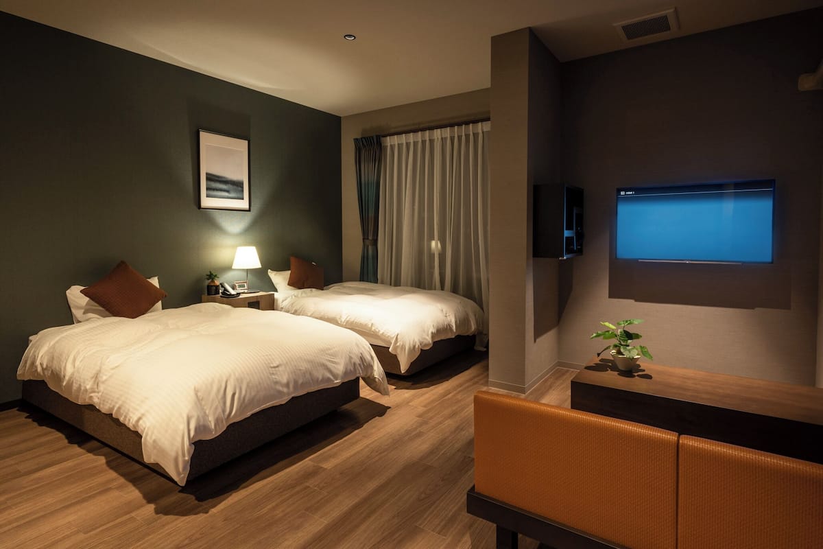 A hotel room with two beds, a desk, a chair, a TV, and a plant.