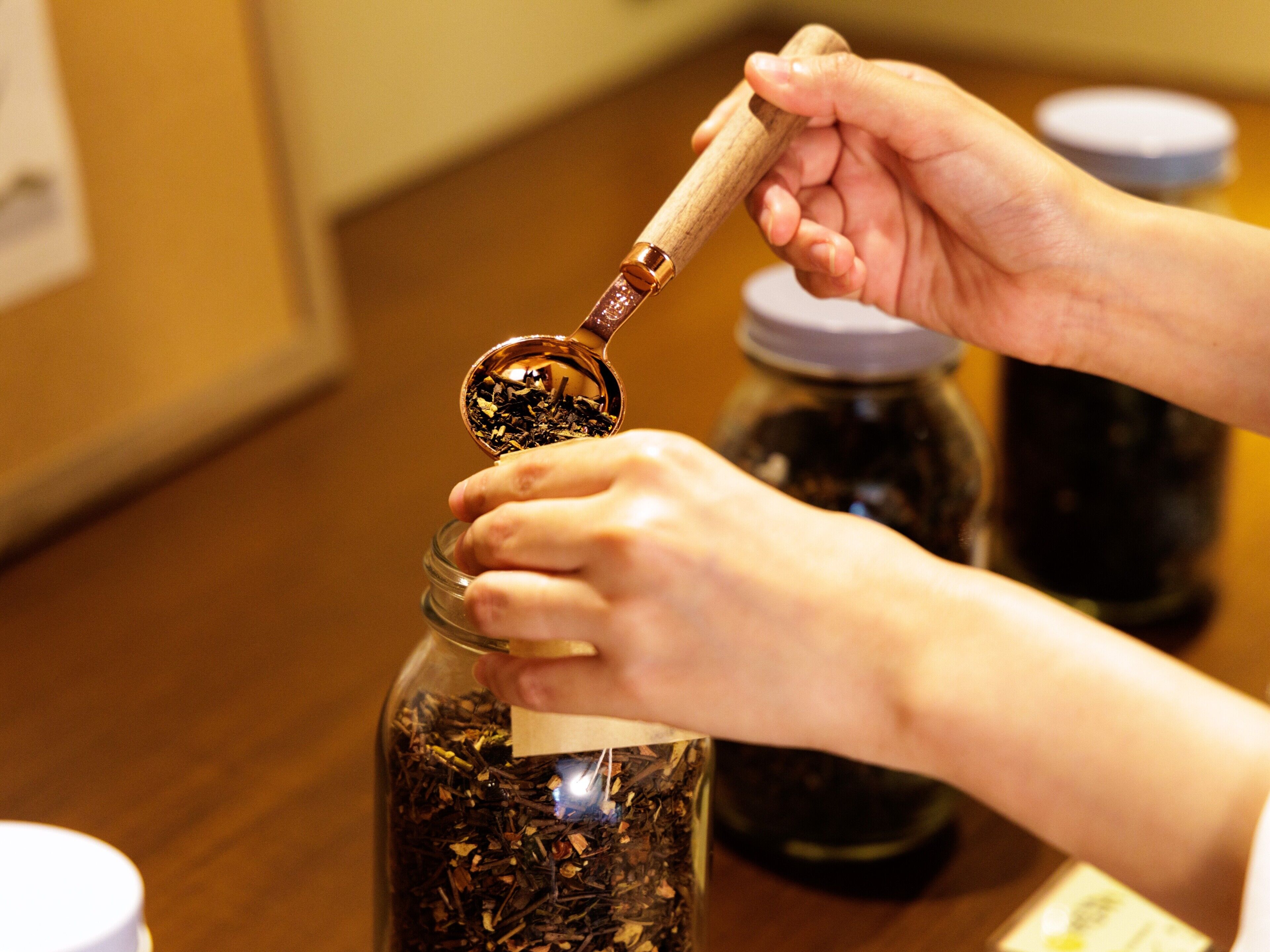 Hands holding a spoon over a jar filled with tea leaves.