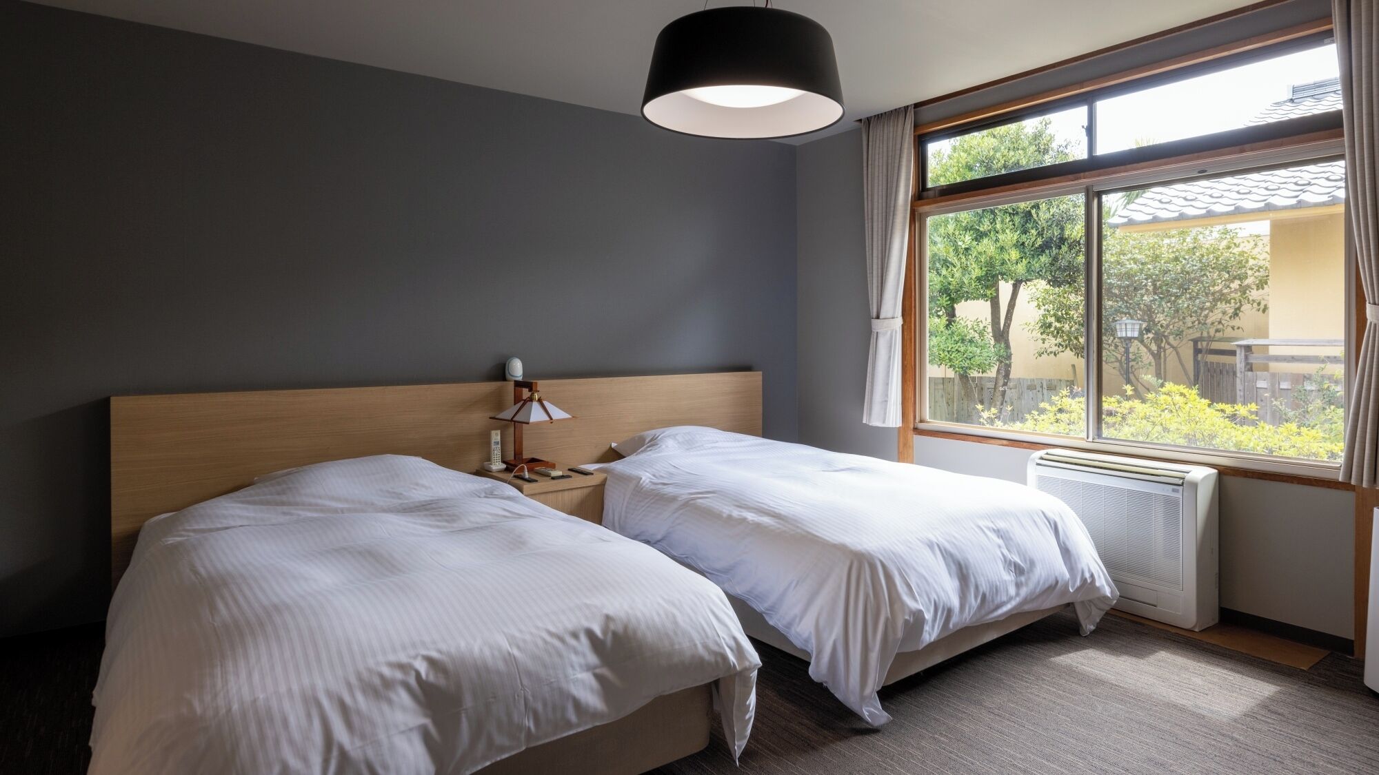 A hotel room with two beds, a bedside table with a lamp, a window with a view of greenery, and a ceiling light fixture.