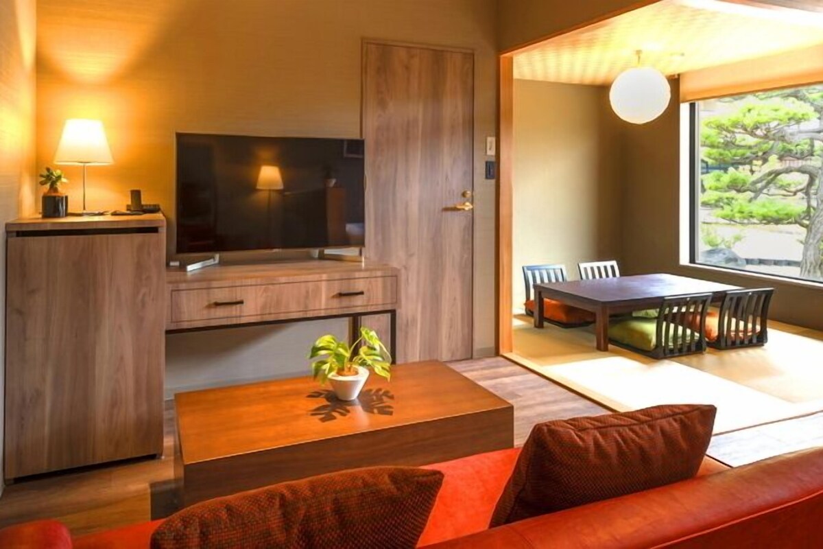 A modern living room with a TV, wooden furniture, a dining area, and a large window with a view.
