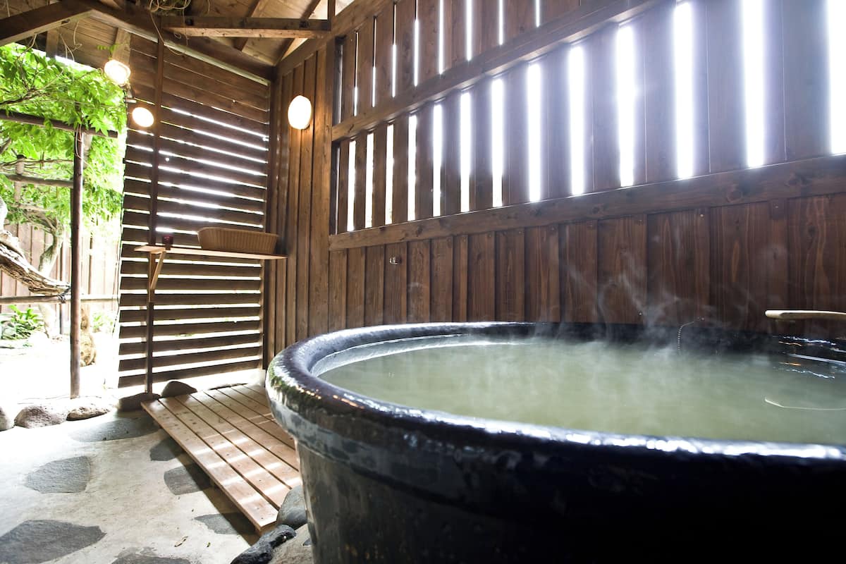 A hot spring bath area with a wooden structure, a large black hot spring tub, and a wooden bench.