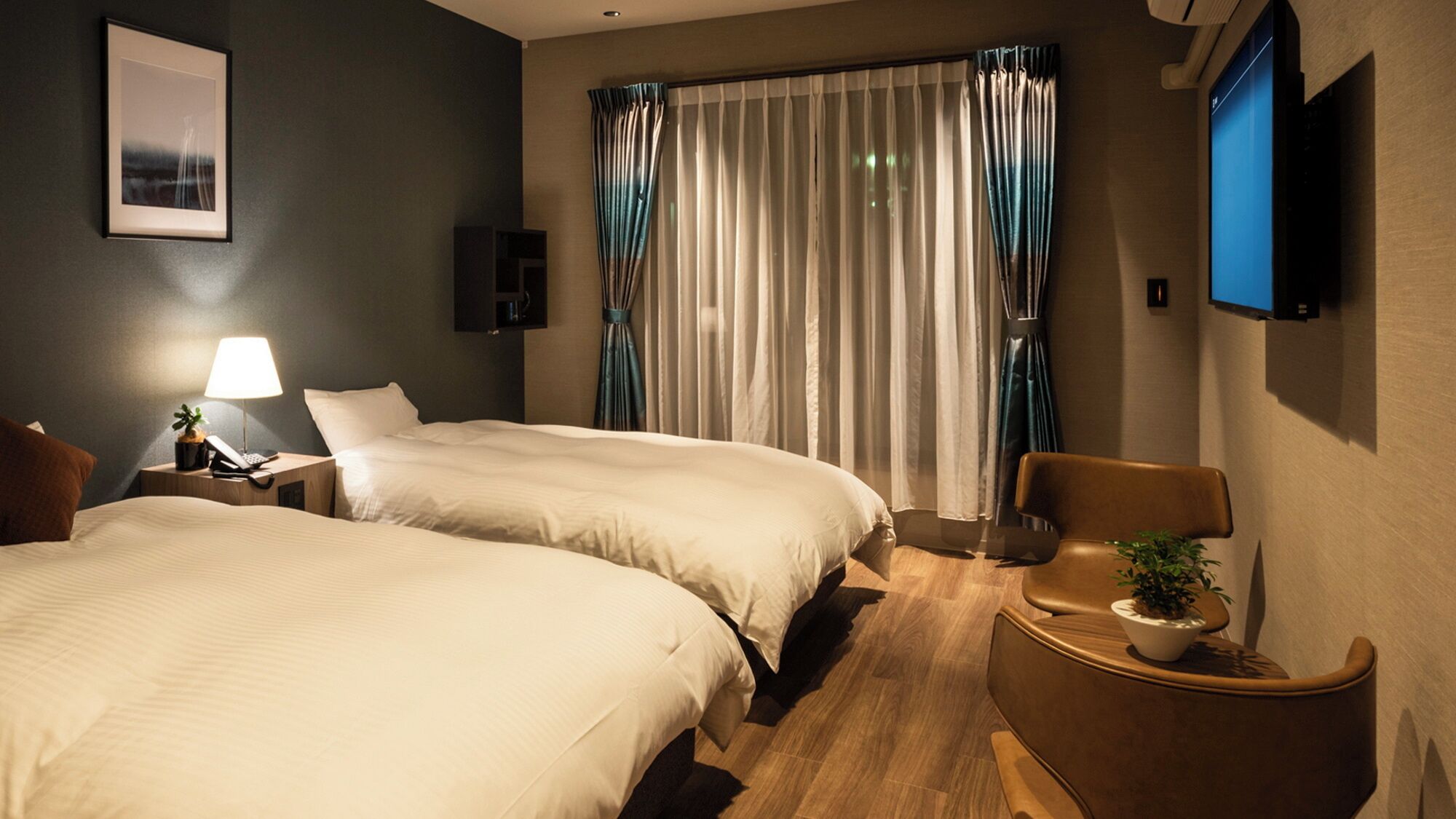 A hotel room with two beds, a desk with a lamp, a chair, a TV mounted on the wall, and a window with curtains.
