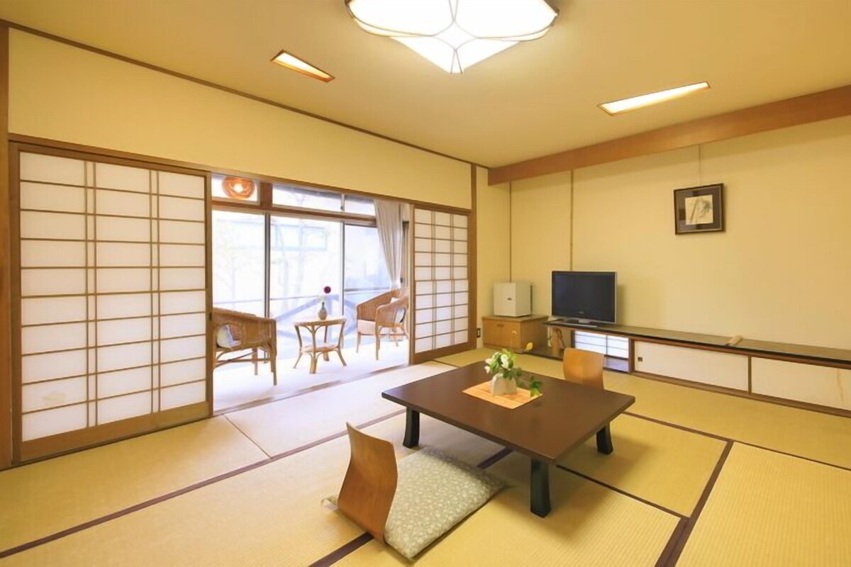 A traditional Japanese room with tatami flooring, a low table with a vase, sliding doors, a television, and a framed picture on the wall.