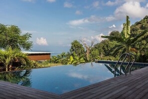 A pool with a wooden deck, surrounded by lush greenery and a view of the ocean.