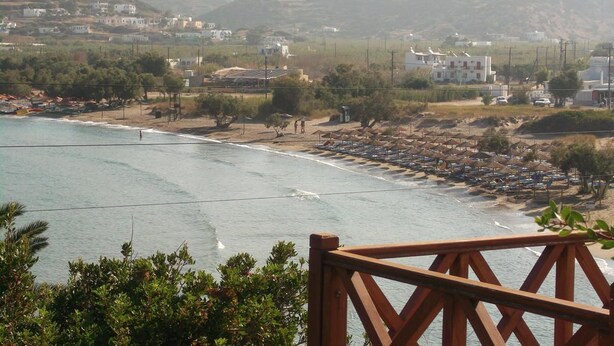 A coastal view with a beach, buildings, and hills in the background. - Manousos' Rooms (Syros)