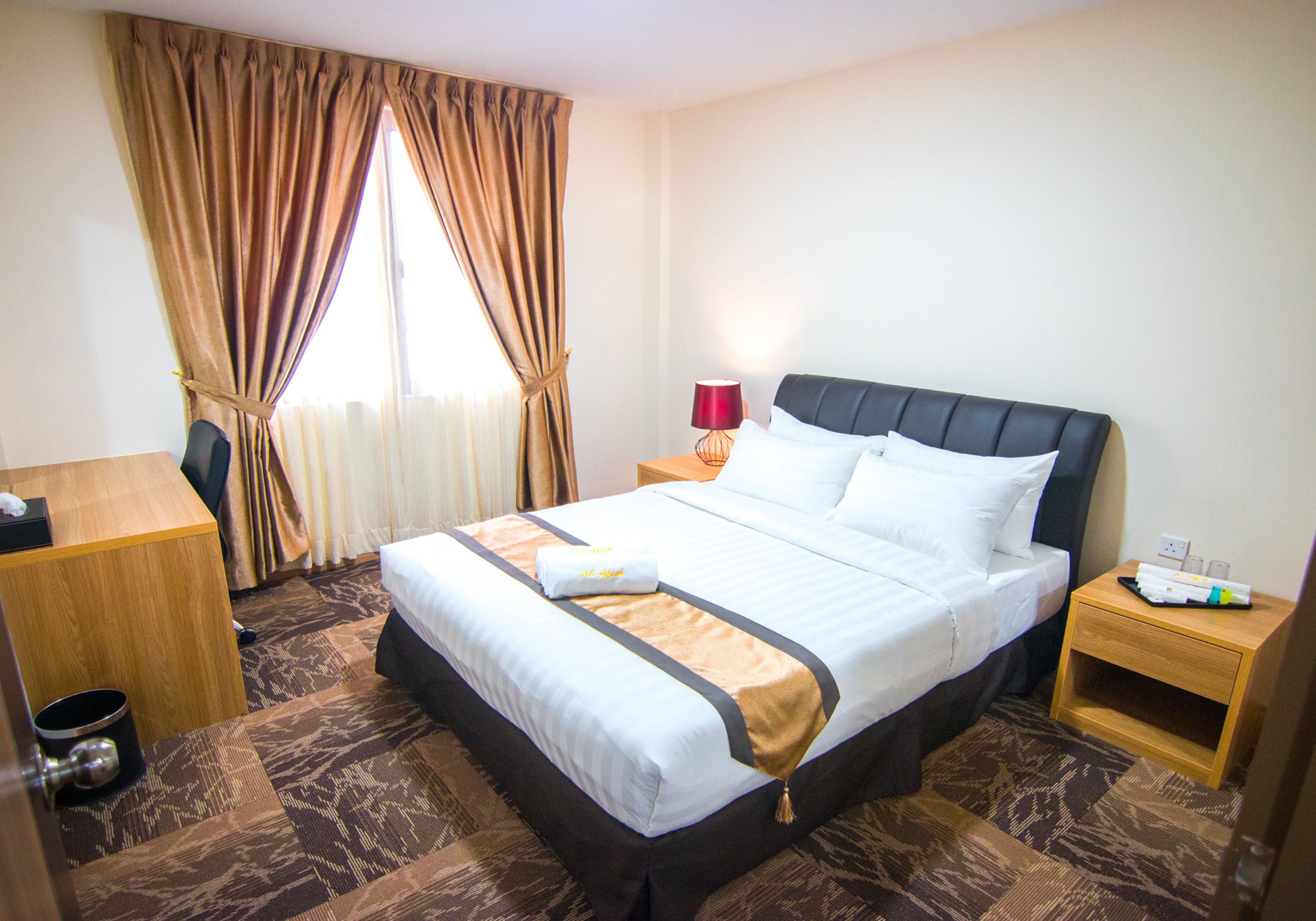 Una habitación de hotel con cama, escritorio, mesita de noche, lámpara y ventana con cortinas.
