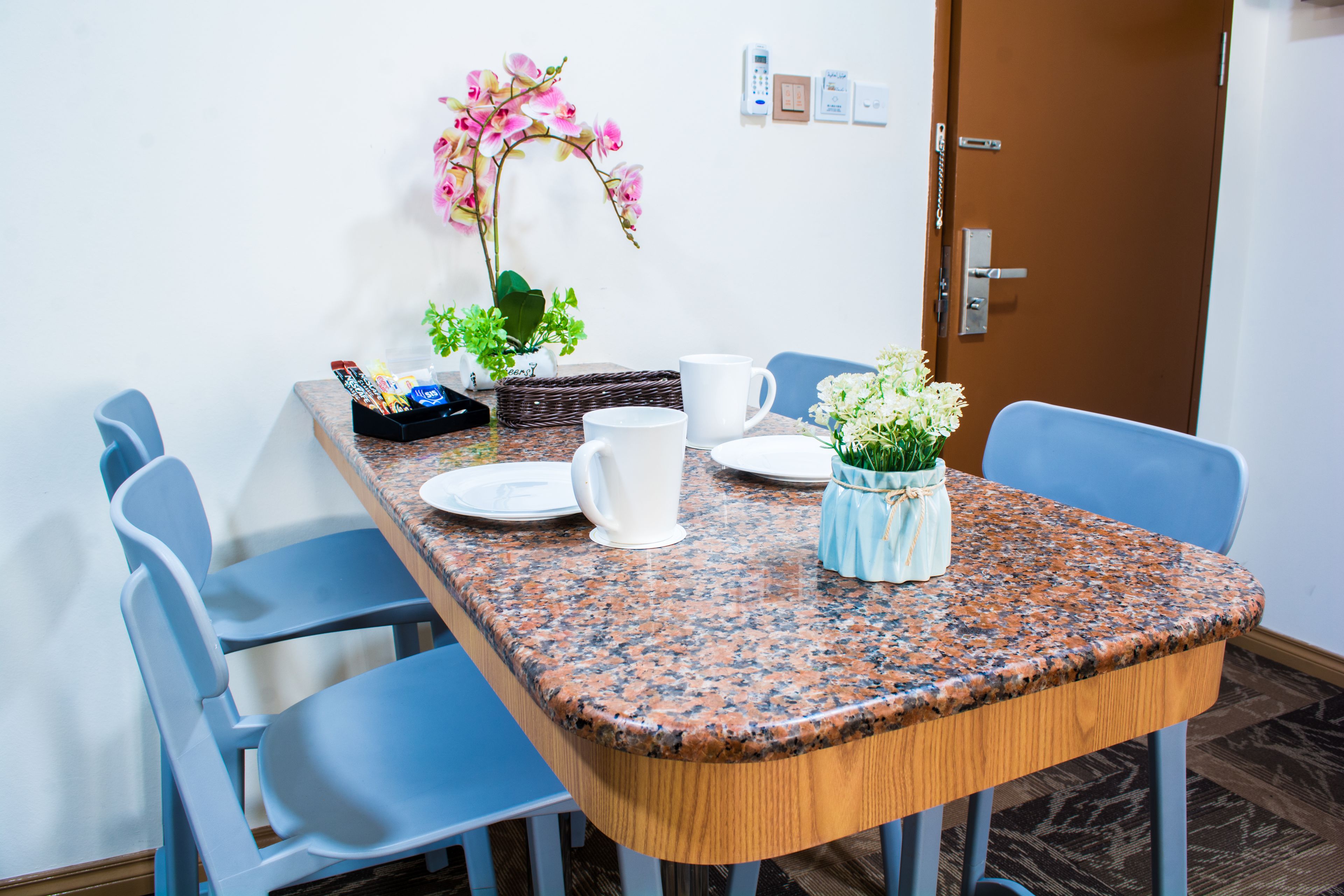 A dining area with a granite table, blue chairs, white plates, a white pitcher, and a small vase with flowers.